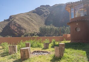 A rustic outdoor area with a fire pit surrounded by wooden stumps, a mountain backdrop, and a building with a circular window. - Bright & Spacious Private Studio w/ Everything! (Písac)