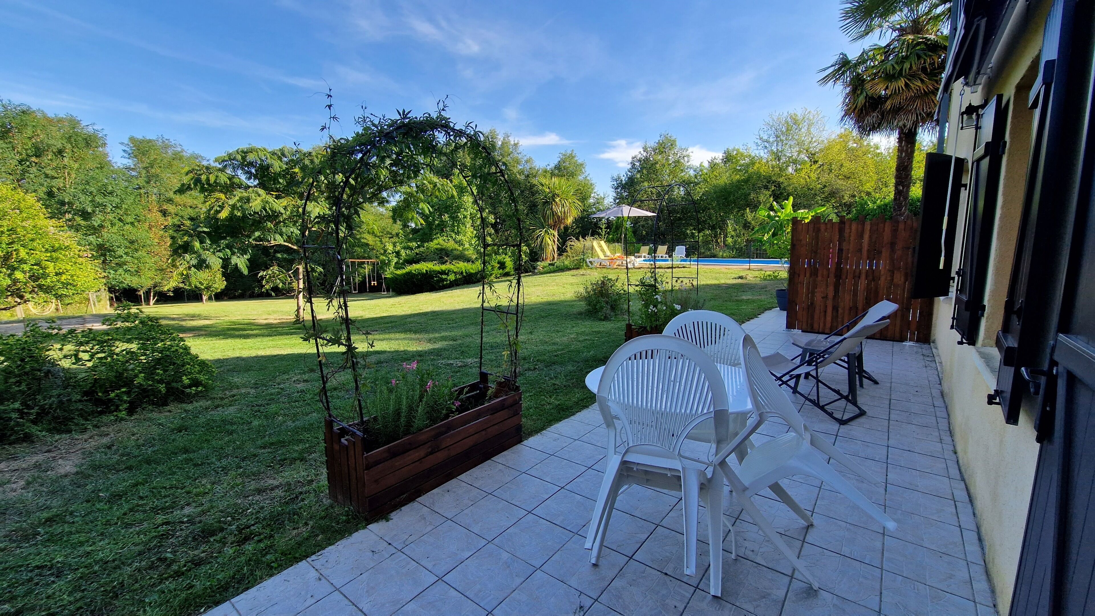 A patio with white plastic chairs and a small table, surrounded by a garden with a swing set and a wooden planter.