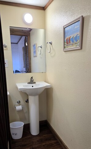 Un baño con lavamanos blanco, un espejo, un cuadro en la pared y un cubo blanco.