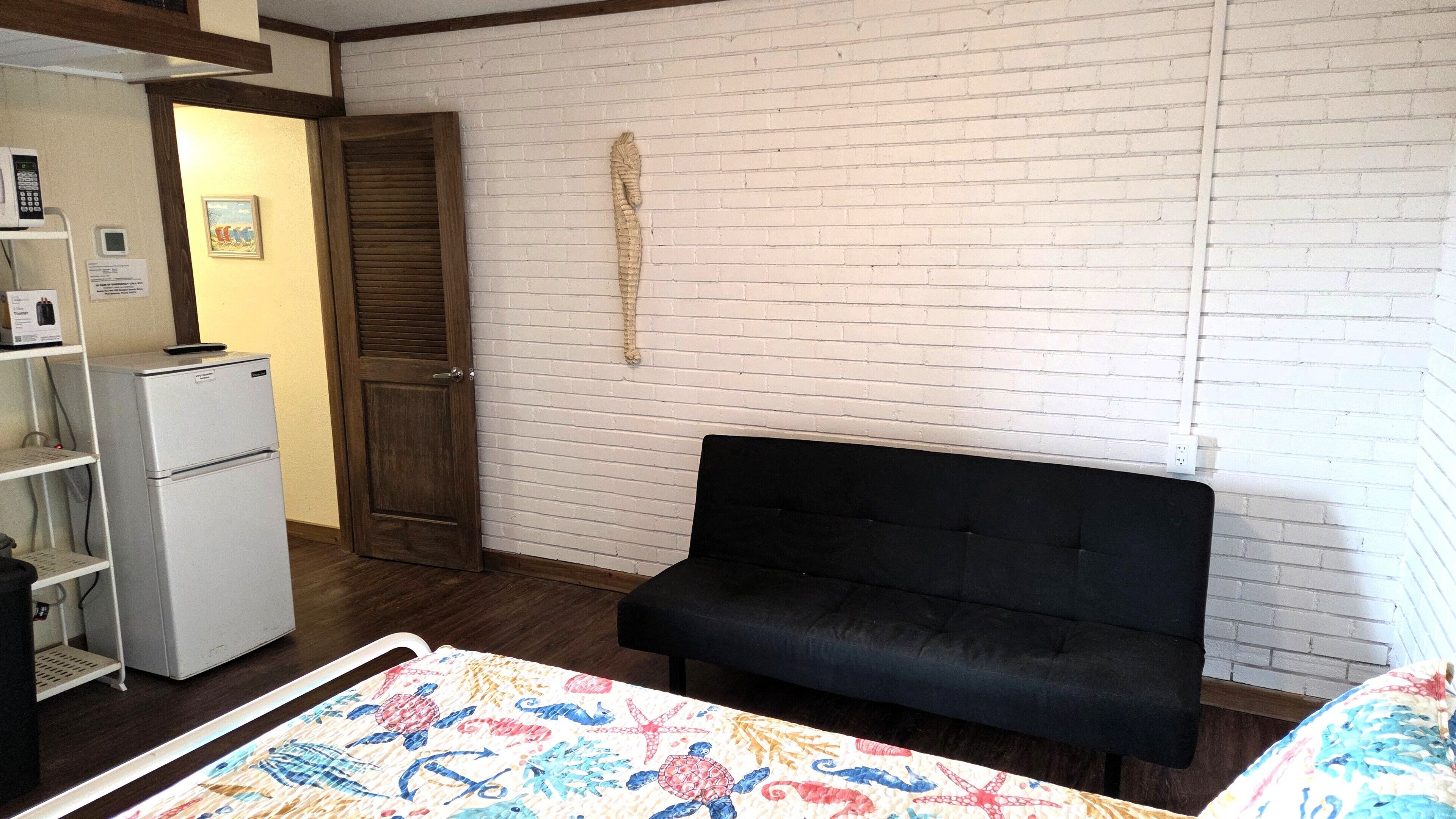 Una habitación con pared de ladrillo blanco, puerta de madera, sofá negro, refrigerador pequeño blanco y colcha de colores en la cama.