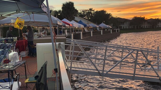 Ein Markt am Wasserufer mit Zelten und Tischen, einem Steg und Blick auf den Sonnenuntergang.