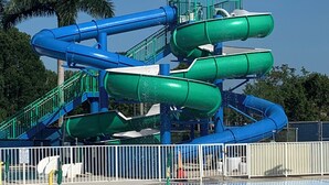 Ein Wasserpark mit mehreren Wasserrutschen in Blau und Grün, umgeben von einem eingezäunten Bereich, im Hintergrund sind Palmen zu sehen.