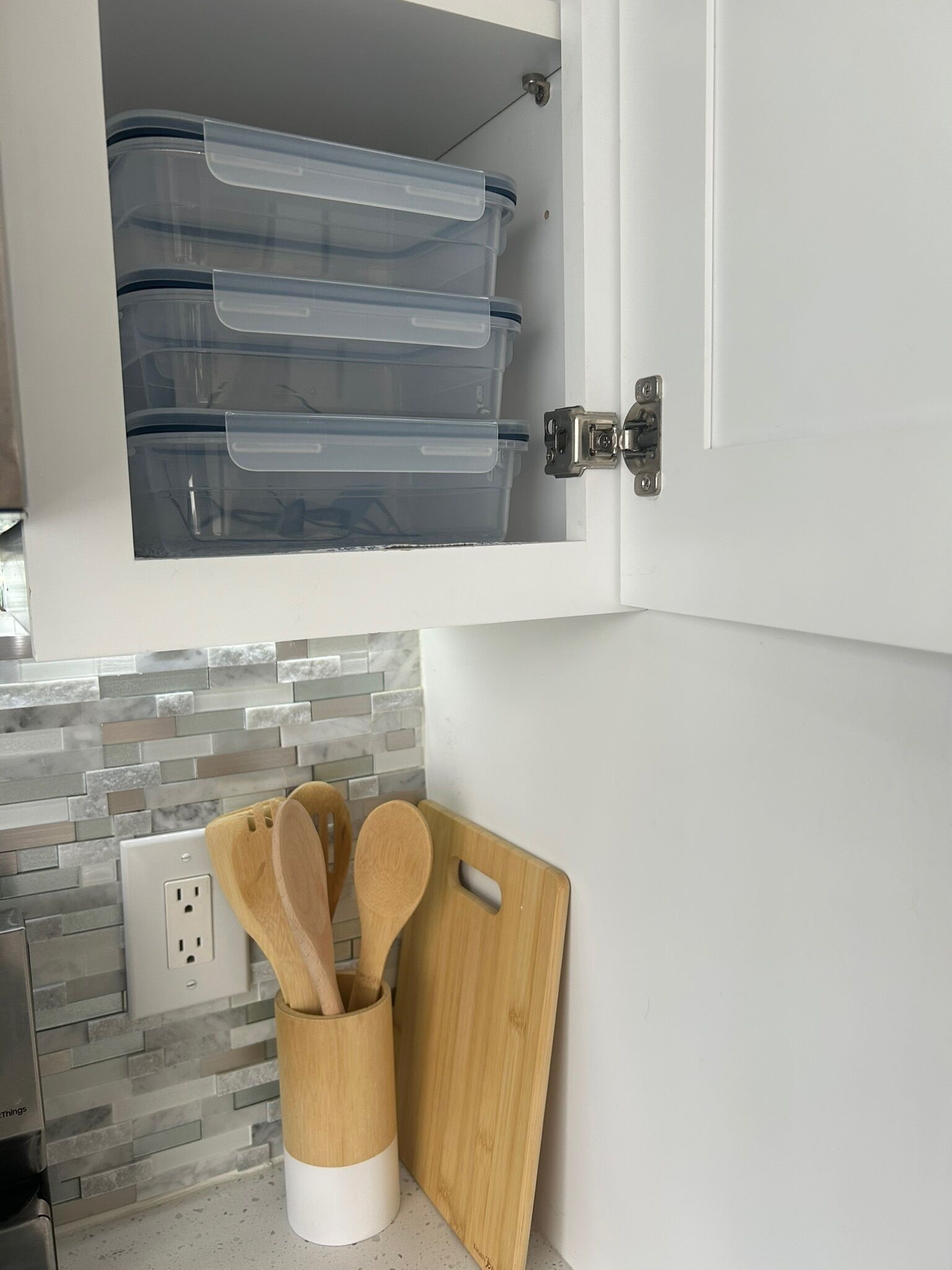 Une cuisine avec un meuble blanc contenant des boîtes de rangement transparentes en plastique, une planche à découper en bois et un porte-ustensiles en bois.