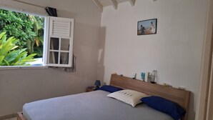 Een slaapkamer met een bed, kussens, een nachtkastje, een raam met luiken en een lijst met een afbeelding aan de muur.