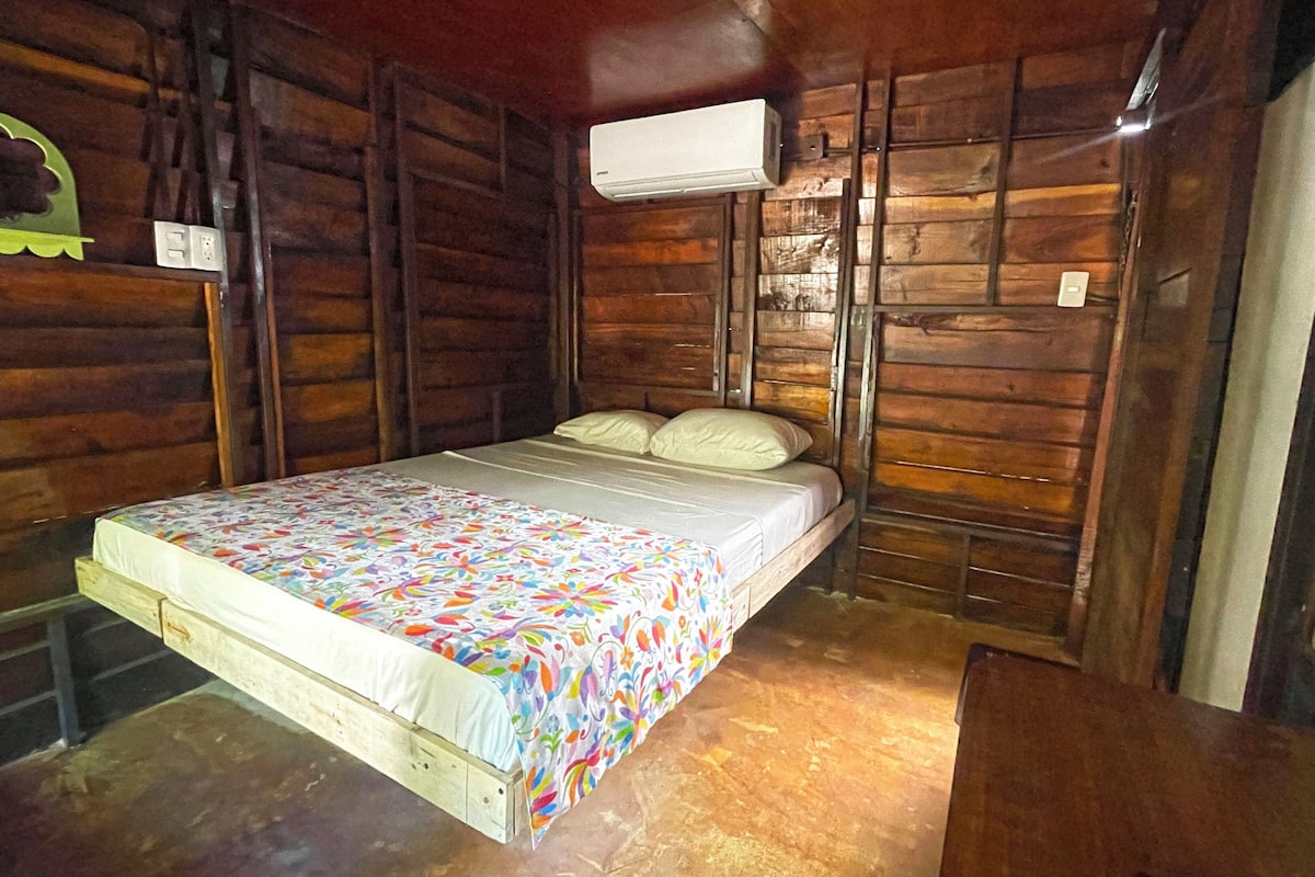 A small, rustic wooden room with a single bed, a wall-mounted air conditioner, and a wooden floor.
