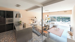A modern kitchen with a built-in oven, a dining area with a glass table and chairs, and a living space with a sofa and a large window offering a view of the outdoors.