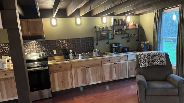 Een moderne keuken met houten kastjes, een inbouwoven en een koffiezetapparaat. Er staat een stoel in de hoek en een raam met gordijnen.