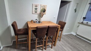 Un comedor con una mesa de madera y cuatro sillas marrones. Hay cuadros colgados en la pared sobre la mesa y un pequeño jarrón con plantas secas.