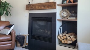 A living room with a fireplace, a brown leather sofa, a grey armchair, a large potted plant, and a wooden shelf with decorative items. - Timberline Retreat - Sleeps 9, large backyard, well stocked kitchen! (Windsor)
