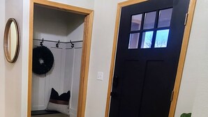 A hallway with a black door, a small seating area with a bench and cushions, a potted plant, and a mirror on the wall. - Timberline Retreat - Sleeps 9, large backyard, well stocked kitchen! (Windsor)