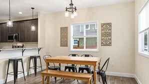 Un espace repas avec une table et des bancs en bois, une cuisine avec des meubles de couleur sombre et des luminaires suspendus.