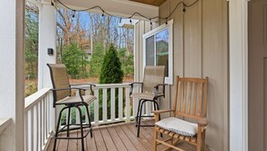 Une véranda en bois avec deux chaises et un banc, une balustrade blanche, et une vue sur des arbres et une maison au loin.