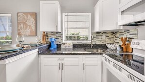Una cocina moderna con armarios blancos, encimera de granito, una zona para café y una ventana con persianas.
