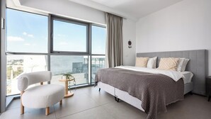 A modern bedroom with a large window, a bed with patterned bedding, a white chair, and a small side table. - MR Savyon view 2BR with 2 balcony 77 (Jerusalem)