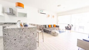 A modern kitchen with a granite island, a grey sofa with orange cushions, and a large window providing natural light. - Seashell Sanctuary -S2-107- 2 bed, 2 bath (Punta Chame)