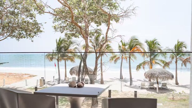 A beach view from a balcony with a table and chairs, palm trees, and a thatched umbrella. - Seashell Sanctuary -S2-107- 2 bed, 2 bath (Punta Chame)