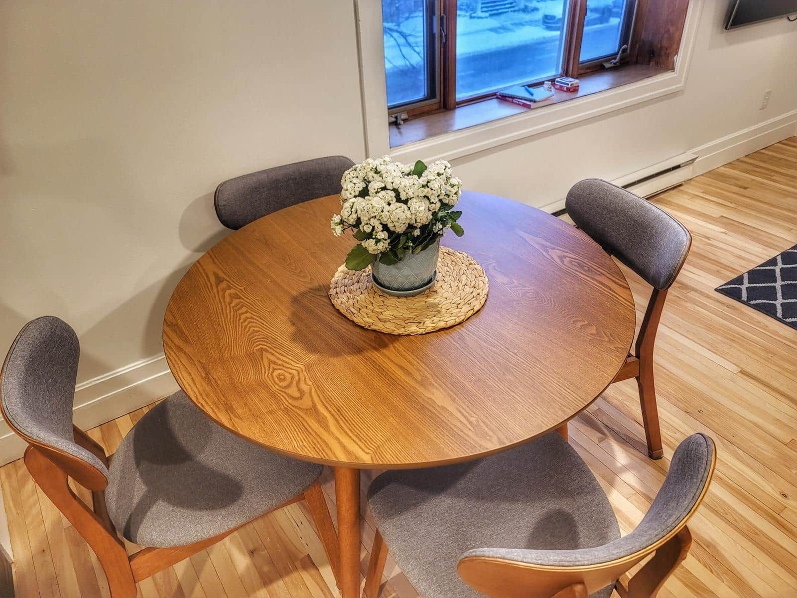 A round wooden table with a woven placemat, surrounded by four grey upholstered chairs.