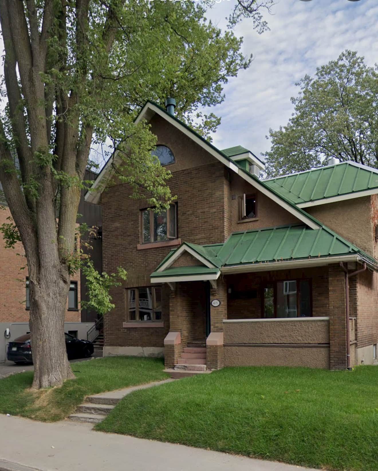 A two-story brick house with a green roof, a front porch, and a large tree in the yard.