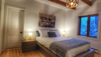 A bedroom with a large bed, wooden beams, a painting of horses, a nightstand, and a window with a view.