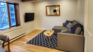 A living room with a sofa, a coffee table, a TV, a mirror, and a window with a view of buildings. - Brilliant 1-bedroom apartment with AC in charming Québec (Québec)