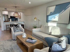A modern living room with a sofa, coffee table, and a dining area with chairs and a kitchen in the background.