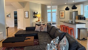 A modern living room with a leather sofa, a wooden coffee table, and a kitchen area with a refrigerator, stove, and orange stools. - Adjacent to UVA Grounds, WALKABLE to Scott Stadium, Corner, Downtown (Charlottesville)