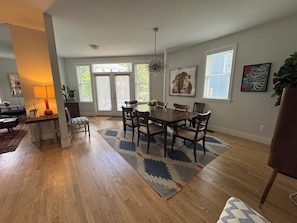 A dining area with a wooden table, chairs, and a patterned rug. There are windows with blinds, a ceiling light, and artwork on the walls.