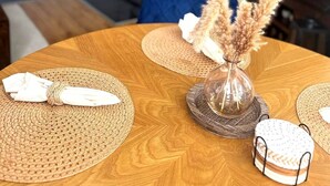 Un comedor con mesa de madera, individuales tejidos y un jarrón con plantas secas.