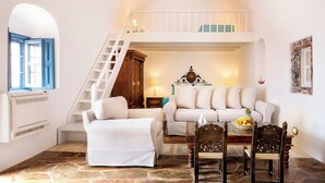 Ein gemütliches Wohnzimmer mit einem weißen Sofa, einem Esstisch mit Stühlen, einer Treppe, die zu einem Schlafboden führt, und einem Fenster mit blauen Fensterläden.