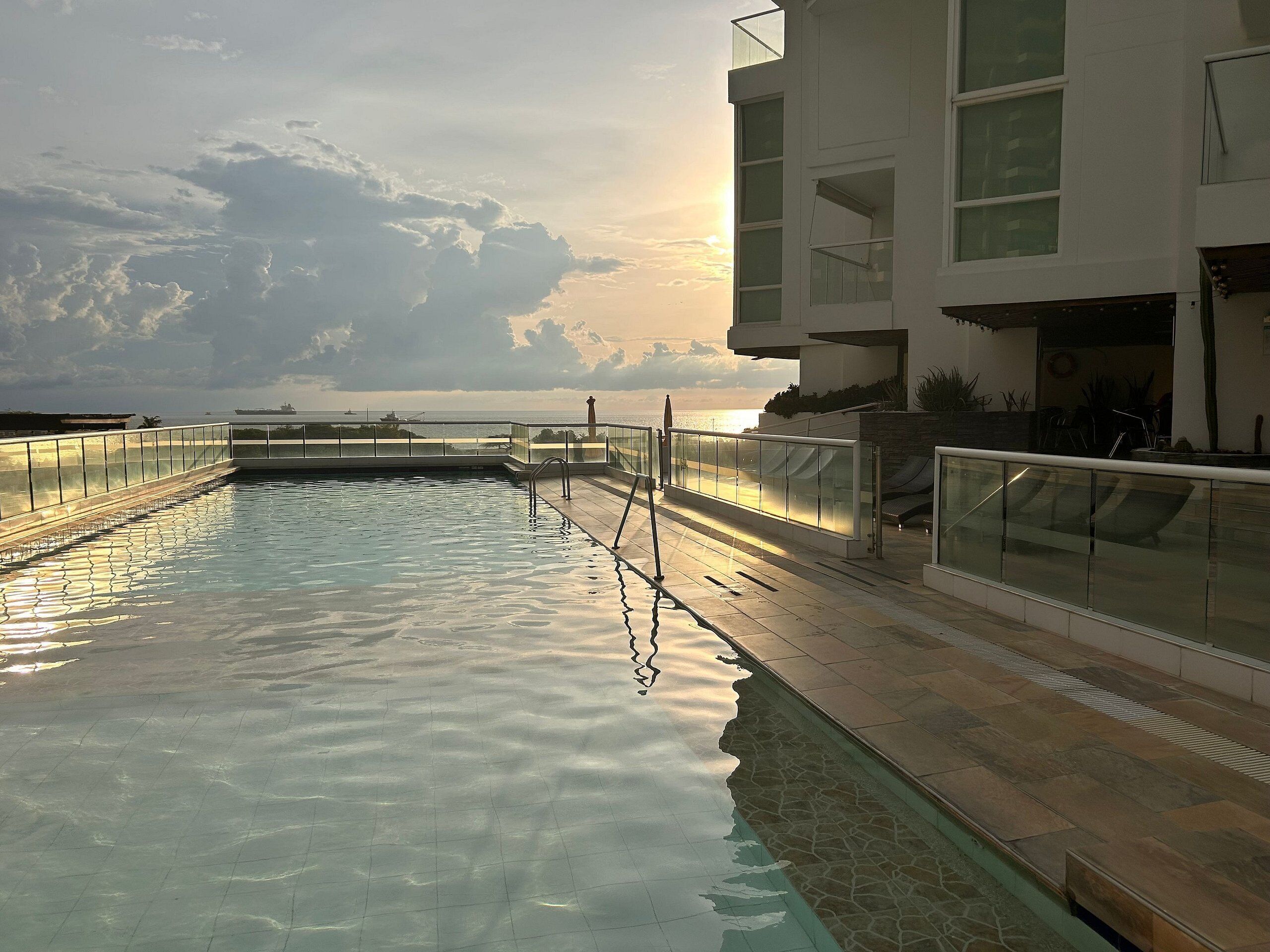 A rooftop swimming pool with a glass balustrade, a view of the sea, and a modern building with balconies.