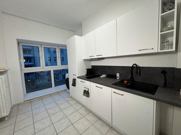 Une cuisine moderne avec des meubles blancs, un plan de travail noir et une fenêtre donnant sur des immeubles.
