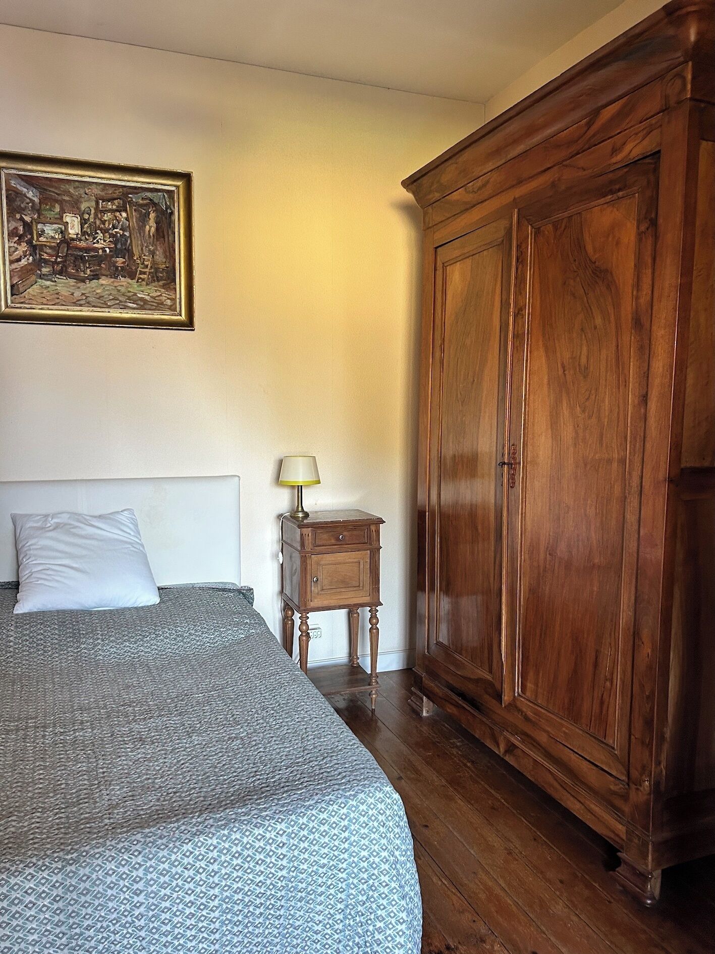 A bedroom with a bed, a wooden wardrobe, a nightstand with a lamp, and a painting on the wall.