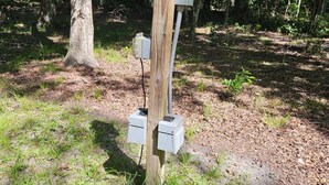 A wooden utility pole with electrical boxes in a forested area.