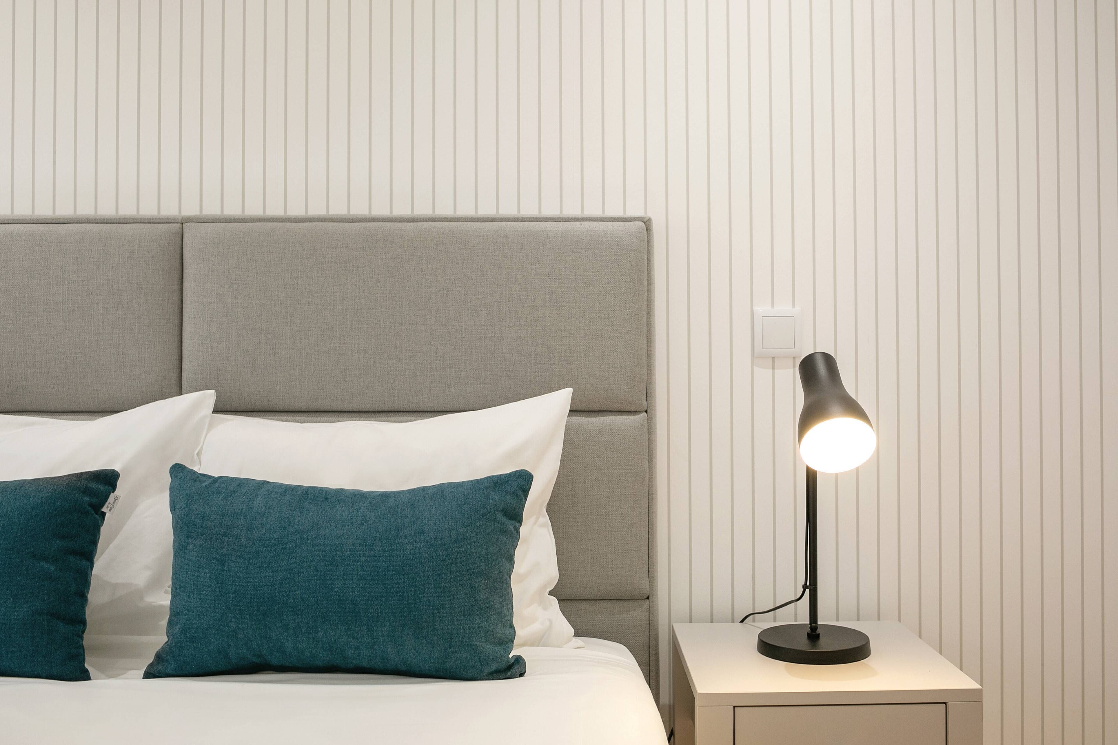 Un lit bien fait, avec une tête de lit grise, des coussins turquoise et une table de chevet surmontée d’une lampe.
