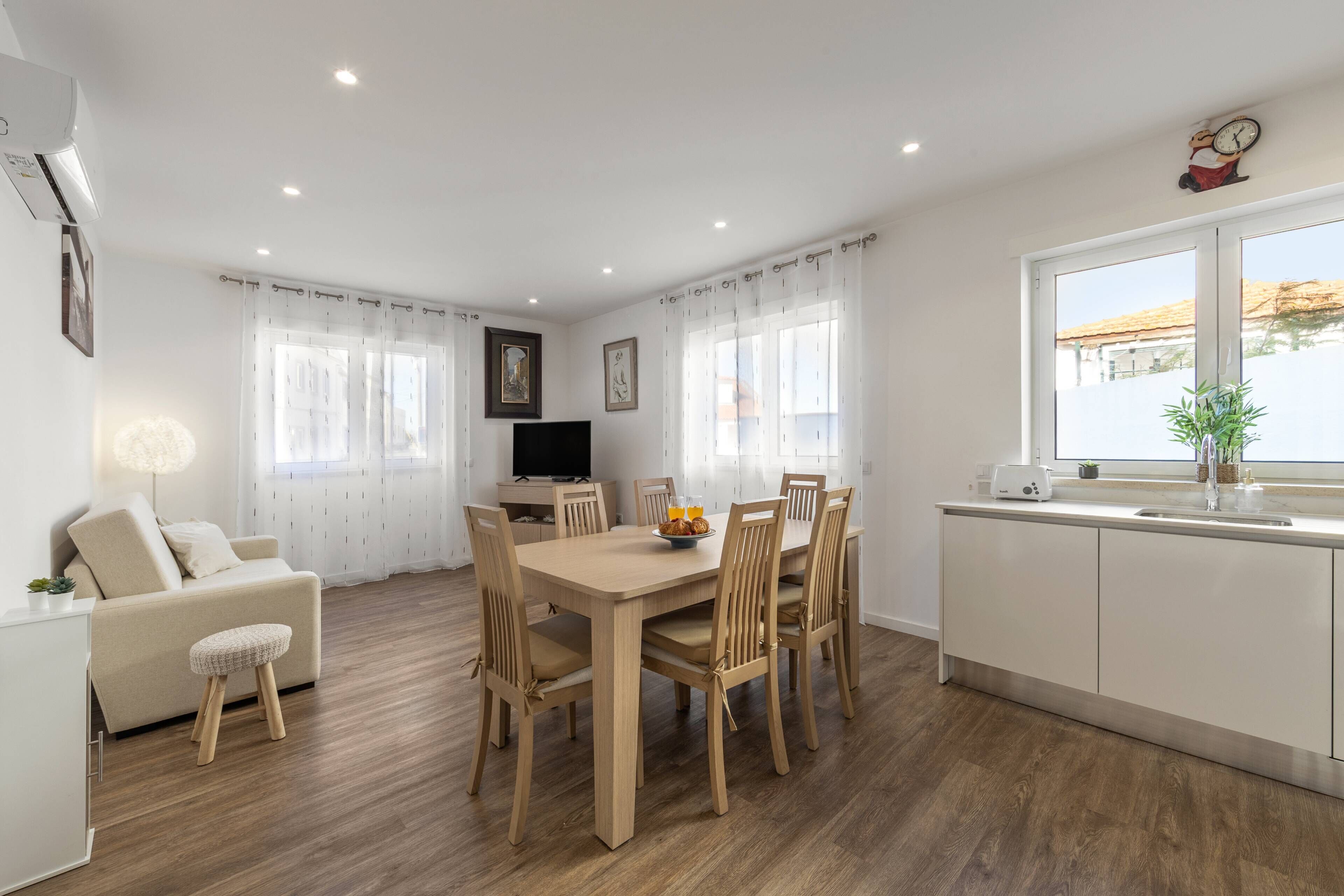 A modern kitchen with a dining area, wooden flooring, white cabinetry, and a large window with curtains.