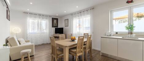 A modern kitchen with a dining area, wooden flooring, white cabinetry, and a large window with curtains.