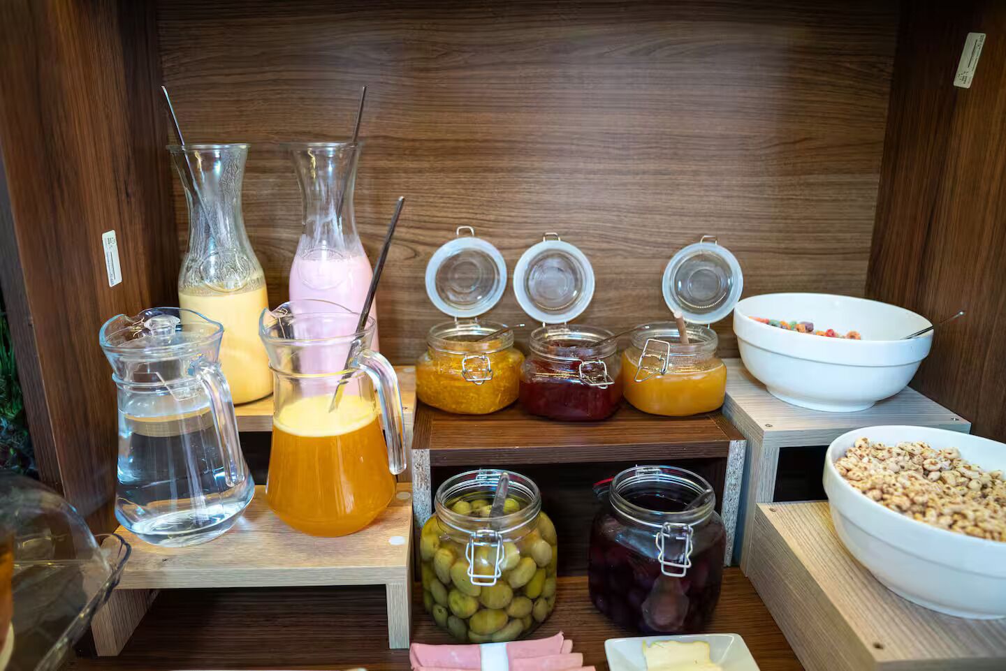 Buffet de café da manhã com diversas opções de comida e bebida, incluindo potes de geleia, um jarro de suco de laranja e uma tigela de cereal.