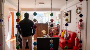 Una persona de pie en un pasillo con decoraciones colgantes de colores, un sofá rojo y un extintor de incendios al fondo.