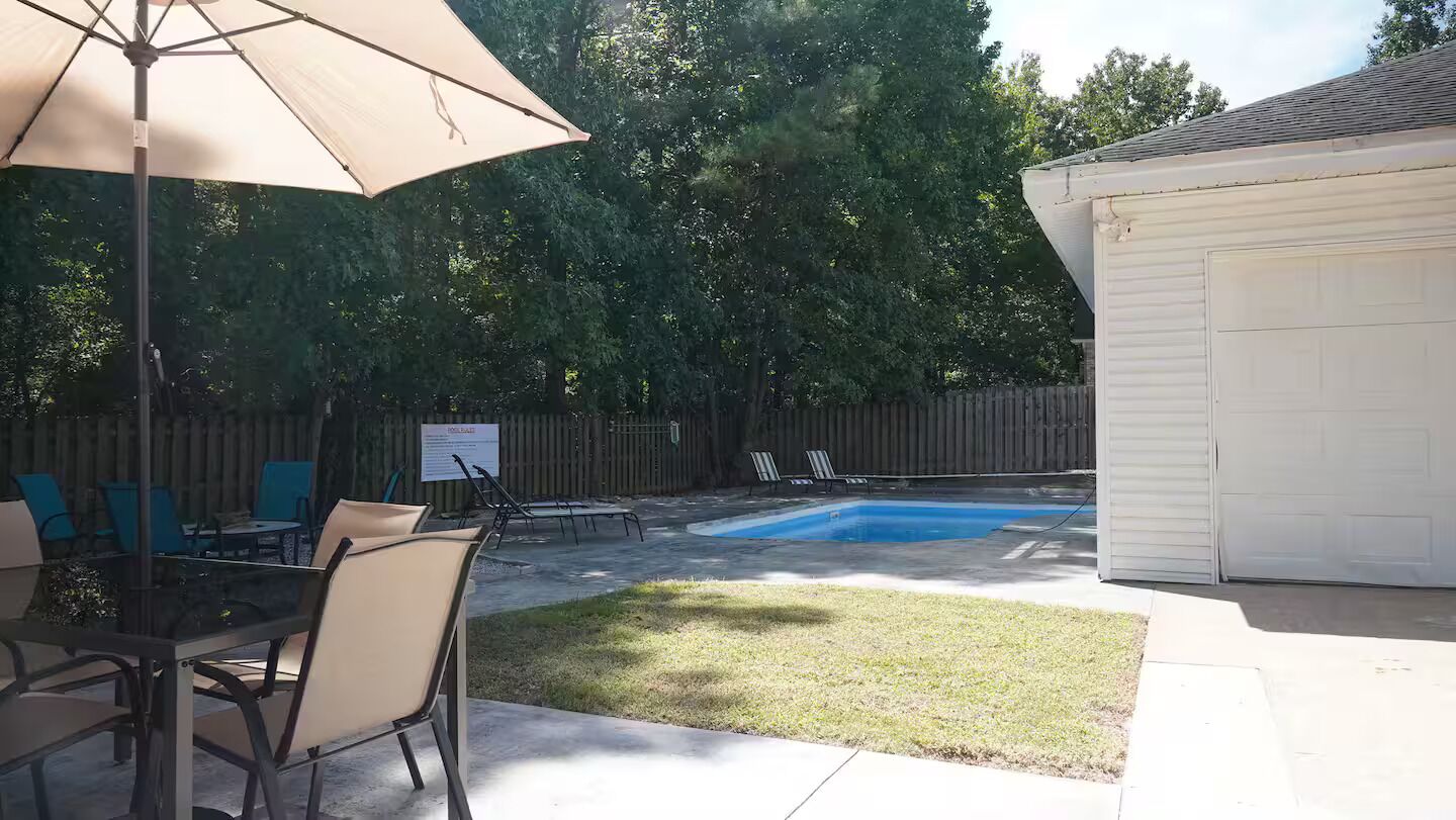 Un patio trasero con piscina, un área de patio techada con sillas y una valla de madera.