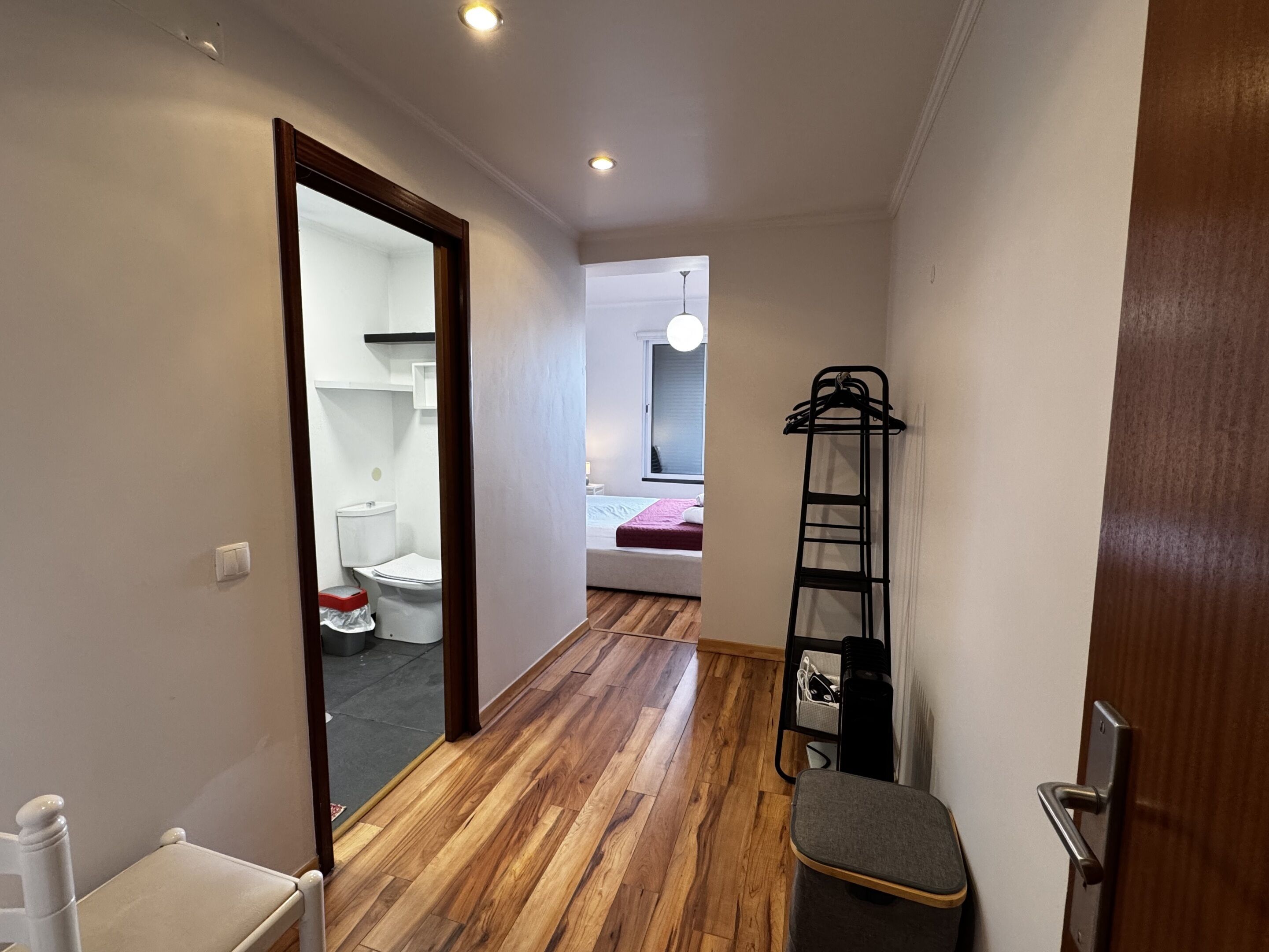 A narrow hallway with wooden flooring, a white chair, a black metal ladder, and a door leading to a bedroom with a bed and a window.