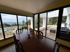 A dining area with a large table and chairs, large windows with a view of the sea and greenery, and a balcony with outdoor furniture.