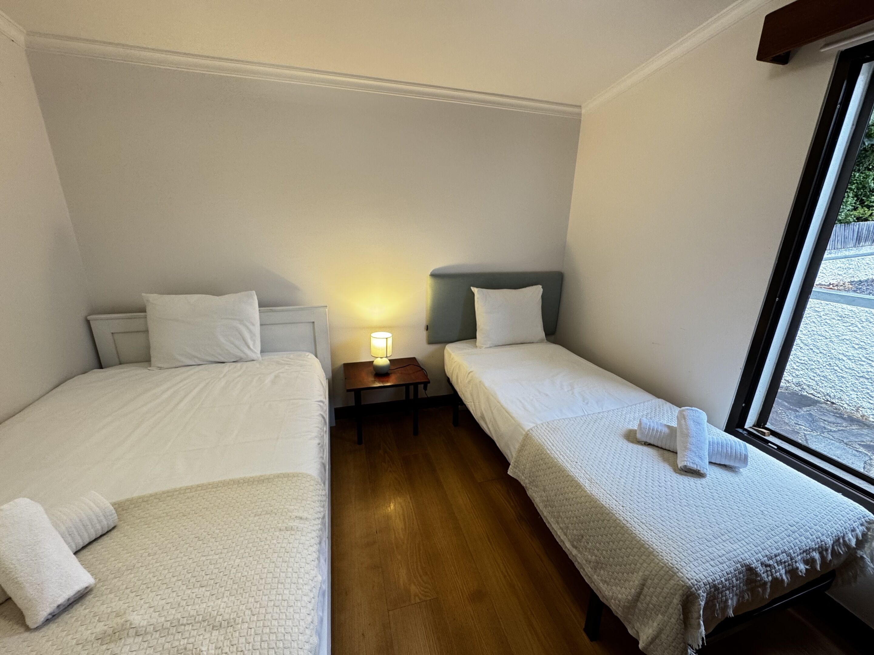 A room with two single beds, a bedside table with a lamp, a window, and a view of the outside.