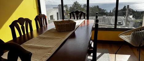 A dining area with a table set for four, a wicker basket, and a view of the sea through large windows.