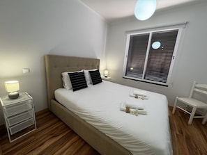 A bedroom with a bed, bedside lamps, a chair, a window with blinds, and a small table with a lamp.