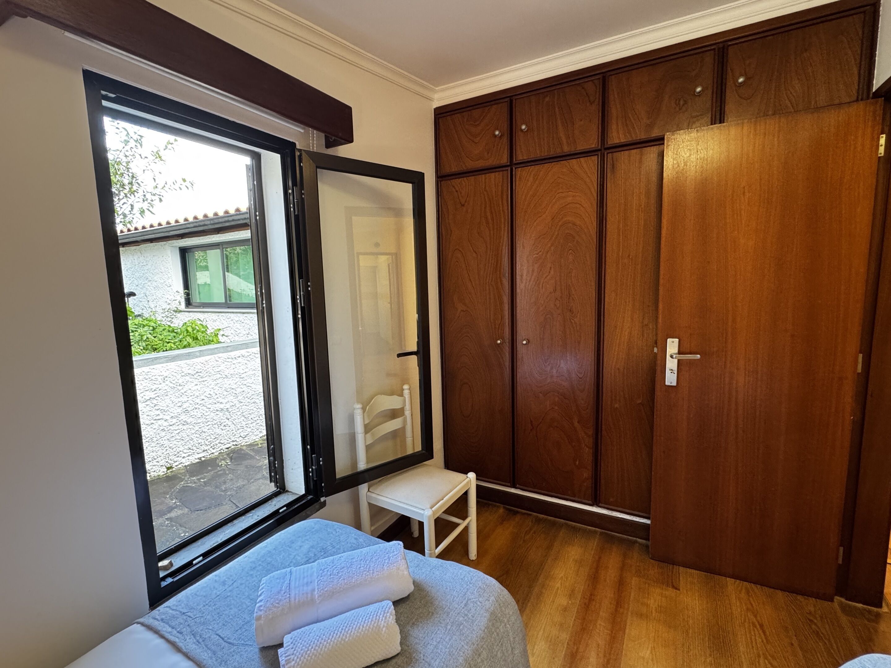 A bedroom with a bed, a wooden wardrobe, a small white chair, and a window.