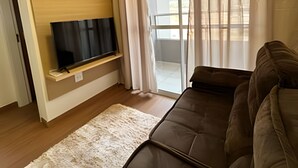 Ein Wohnzimmer mit Flachbildfernseher auf einer Holzablage, einem braunen Sofa, einem weißen Teppich und einer Glastür, die auf einen Balkon führt.