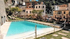 A swimming pool with a wooden deck and lounge chairs, surrounded by terraced buildings and a mountainous backdrop.