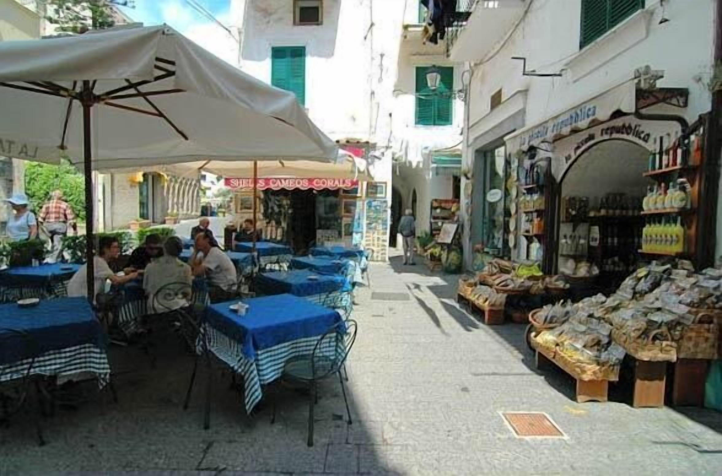 Un café al aire libre con mesas y sillas, una sombrilla blanca, y una calle con tiendas y balcones.