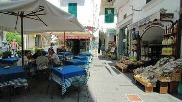 Un café al aire libre con mesas y sillas, una sombrilla blanca, y una calle con tiendas y balcones.