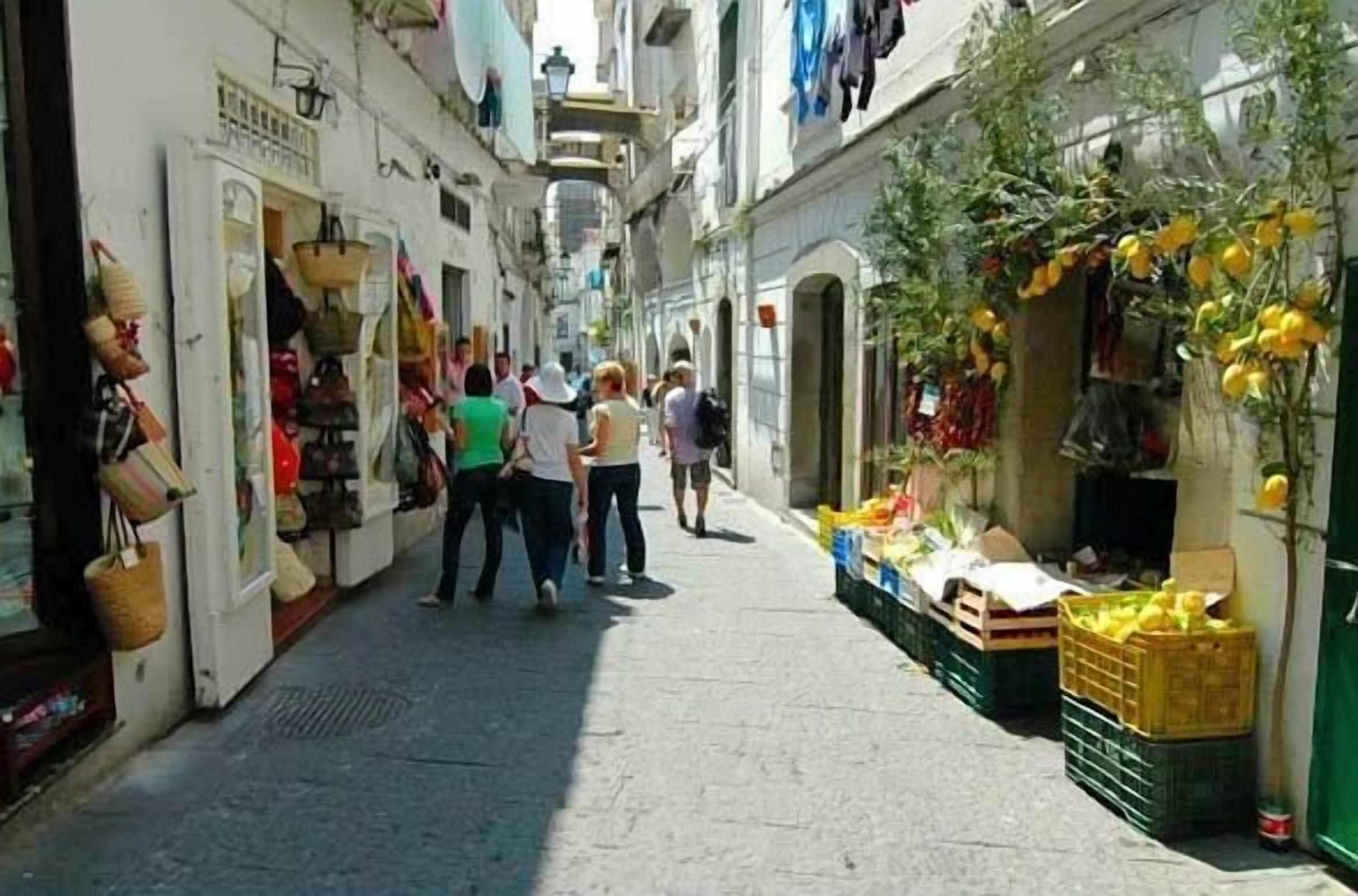 A narrow street with shops on either side, including a fruit stand with yellow fruits.